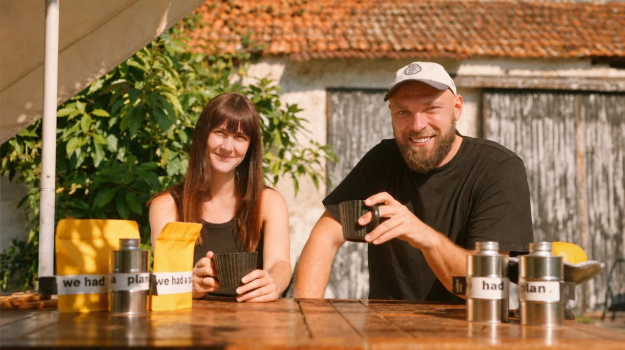 Vadim Key and family at a sunlit wooden table outside an old farmhouse, holding cups, with the yellow bags and silver tins in front of them.